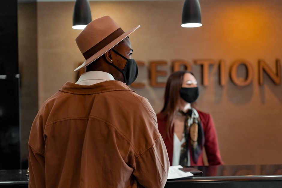 Economia do Turismo — Man in mask at hotel reception during pandemic, interacting with masked receptionist.