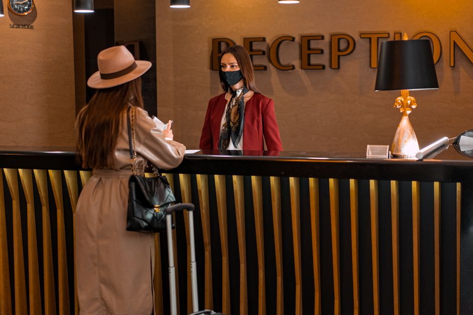 Economia do Turismo — A woman speaks to a receptionist wearing a mask at a hotel reception.