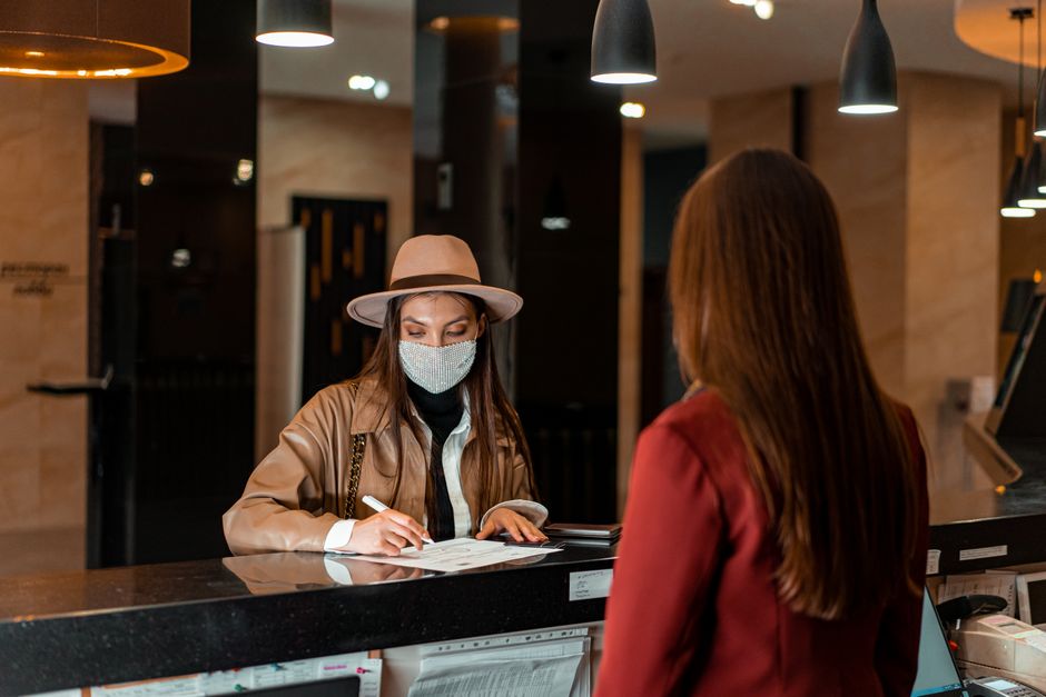Economia do Turismo — A woman in a hat and face mask checks in at a hotel reception, interacting with a receptionist.