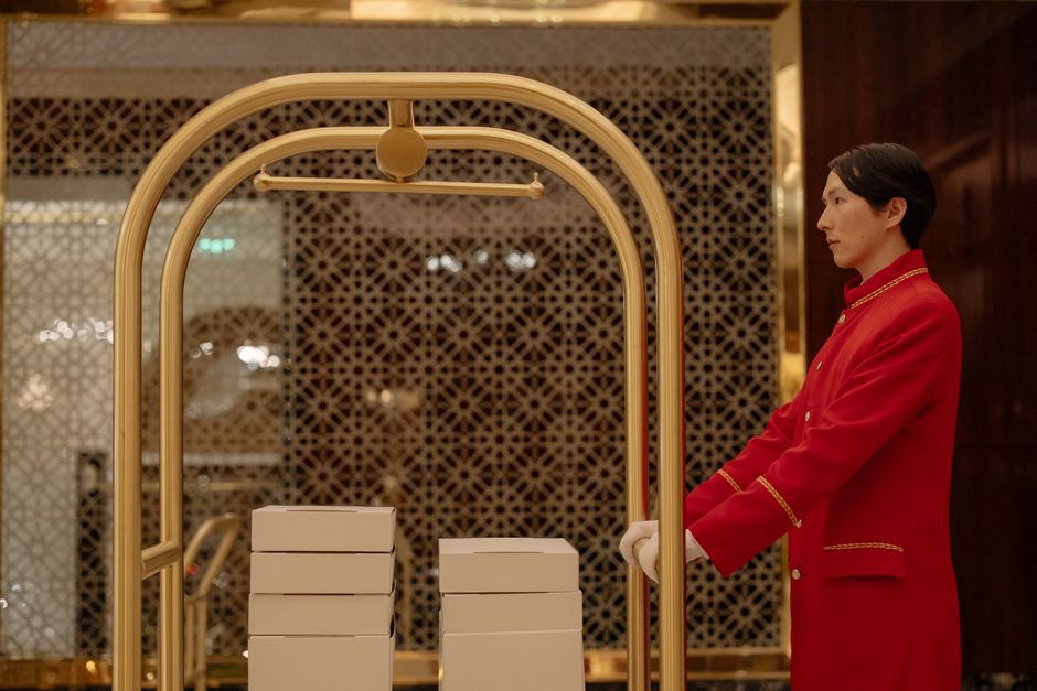 Economia do Turismo — A hotel bellboy in uniform pushes a luggage cart with boxes indoors, demonstrating hospitality service.